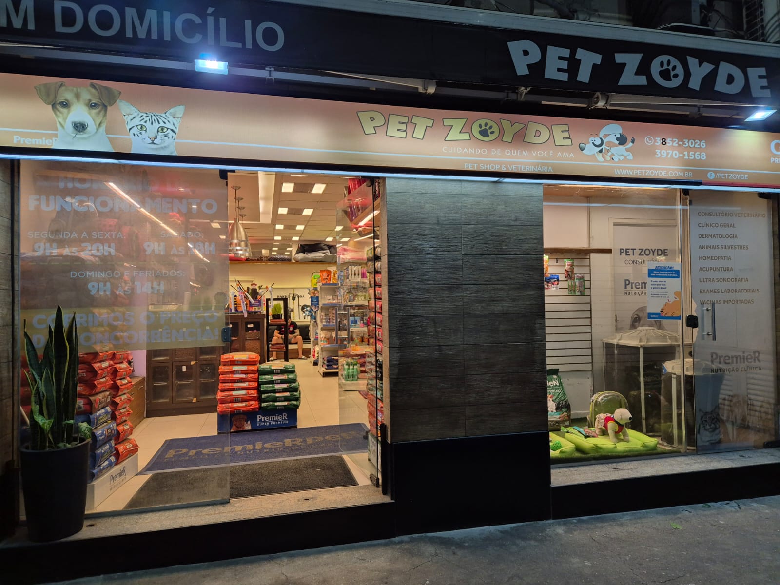 Fachada da Pet Zoyde iluminada à noite, com letreiro Pet Zoyde, vitrine de rações Premier e horário de funcionamento